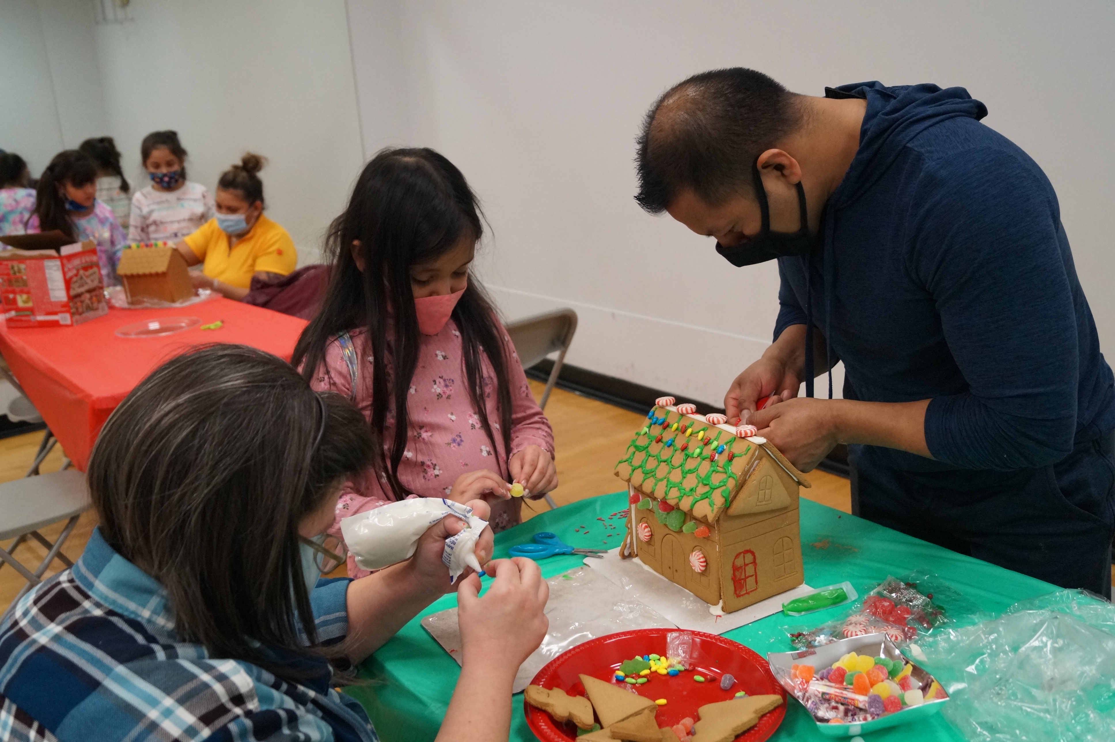 A mother, father, and daughter decorating a gingerbread house with candy and icing.