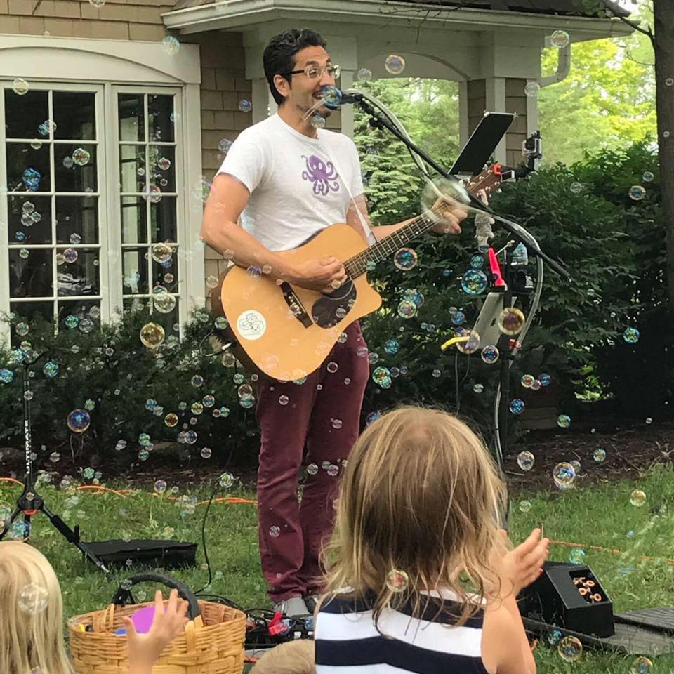 Istvan singing outside while playing guitar. Kids clapping and popping bubbles.