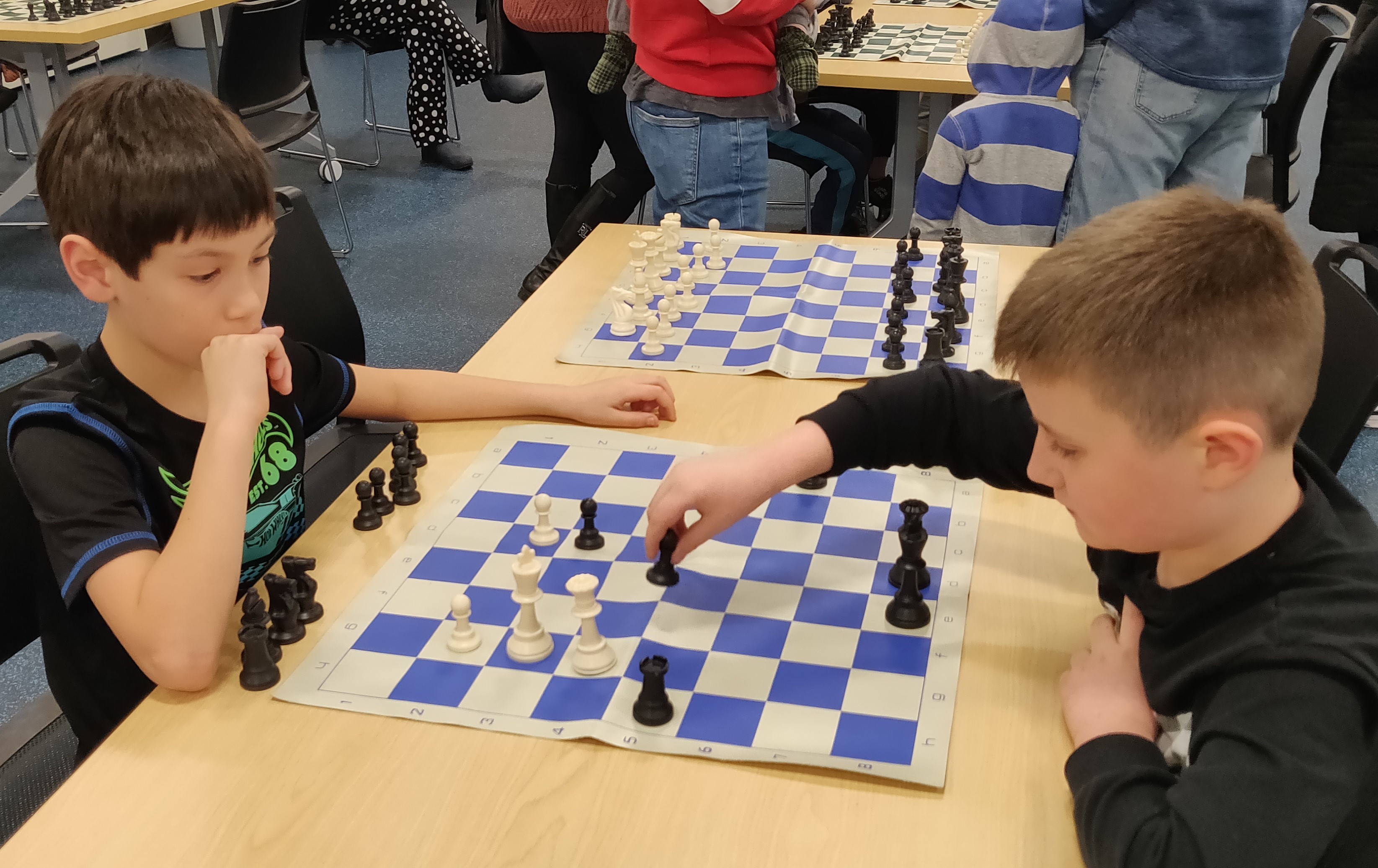 Two kids playing chess. One is moving a piece and the other is thinking about their next move.