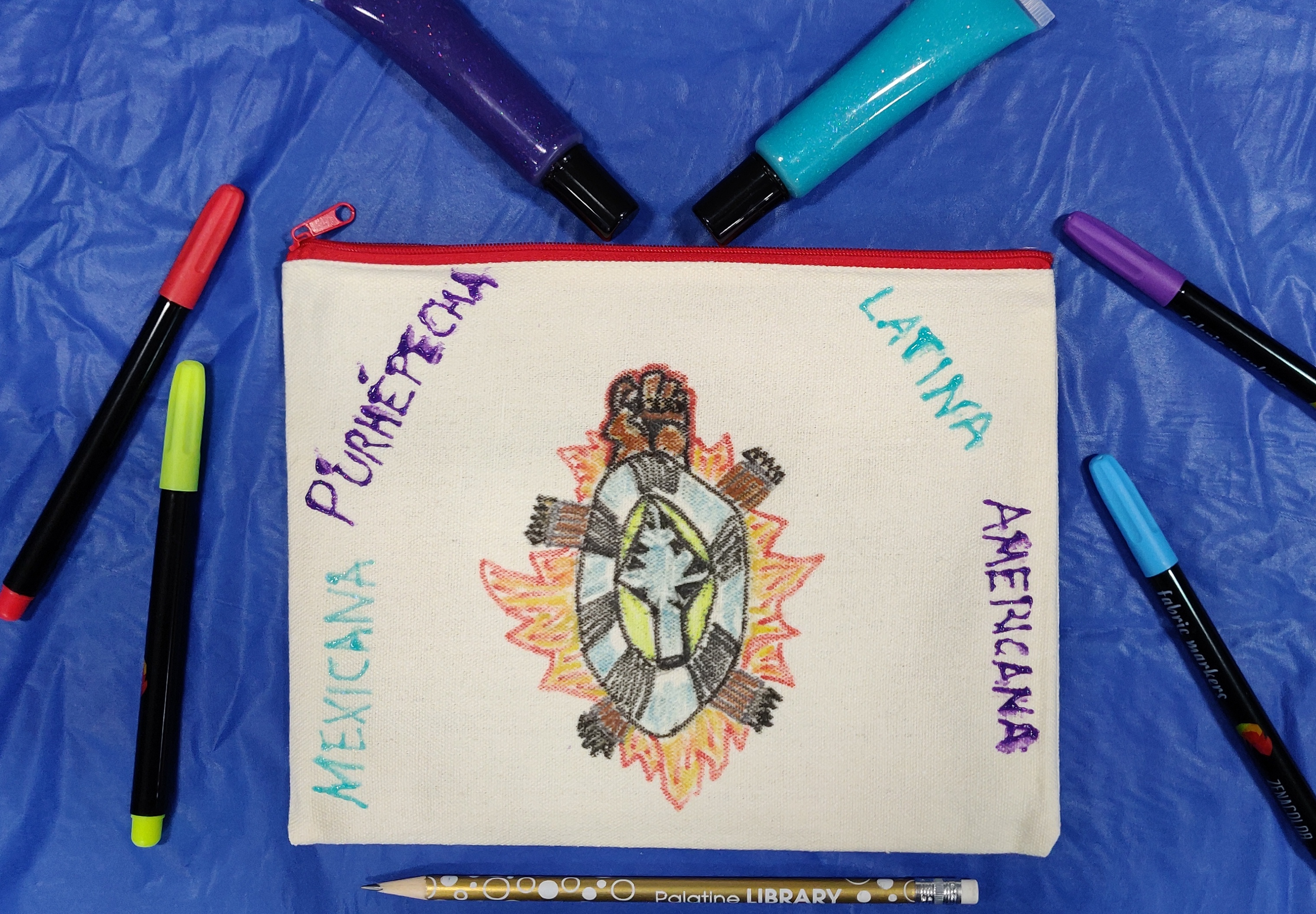 Canvas bag that has been drawn on surrounded by fabric markers, a pencil, and fabric paint. 