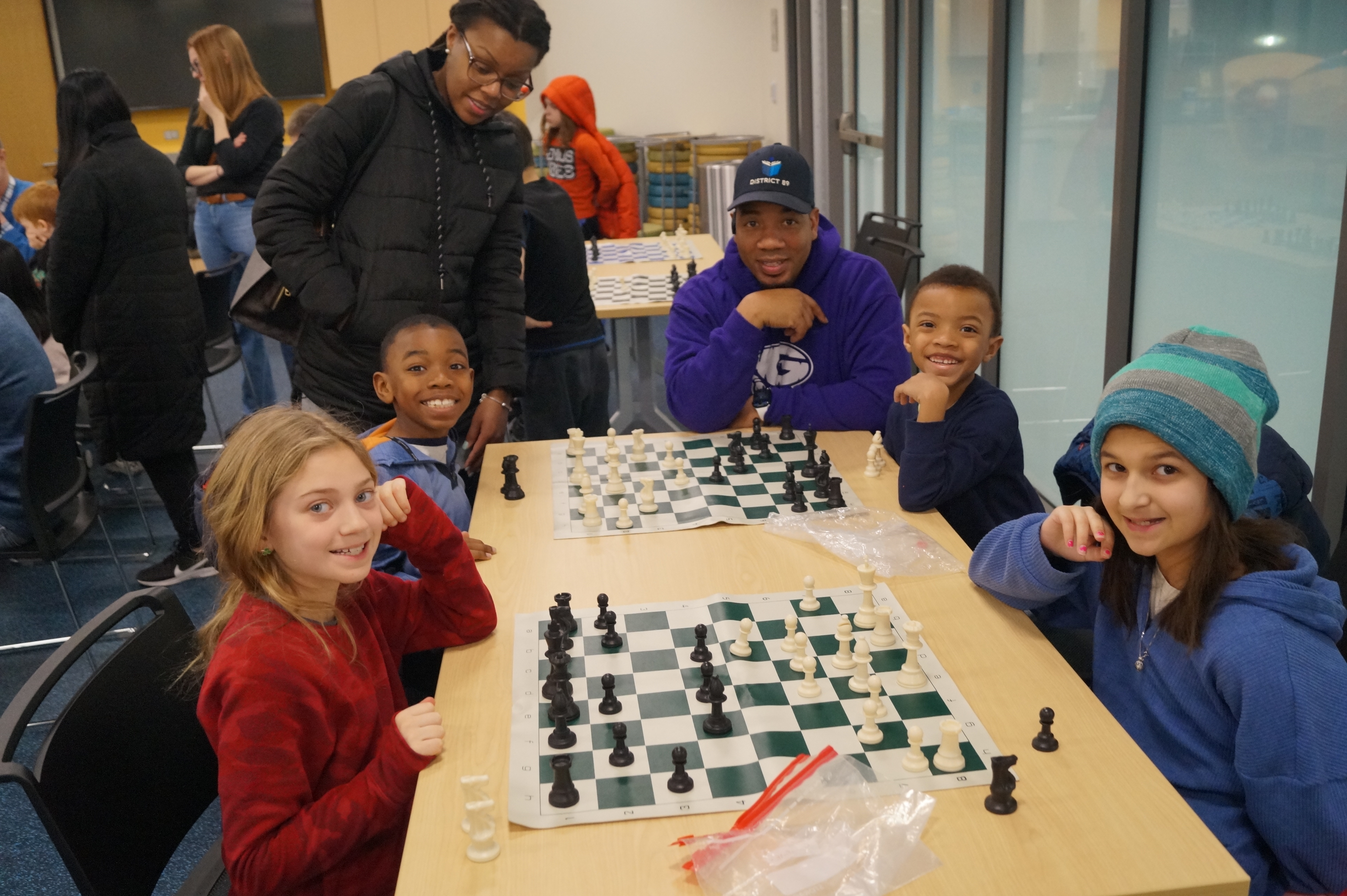 Four kids in front of chess sets smiling at the camera. Two parents alongside their children.