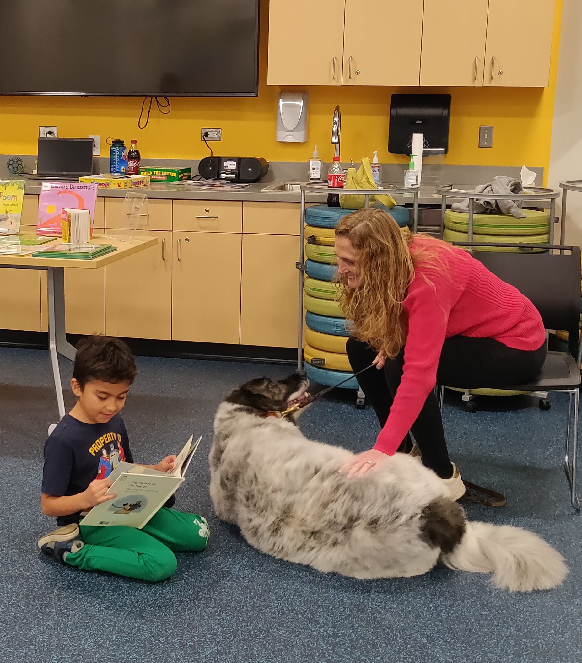 A little boy reading a picture book to a dog while the dog's handler smiles.