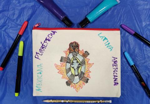 Canvas bag that has been drawn on surrounded by fabric markers, a pencil, and fabric paint. 
