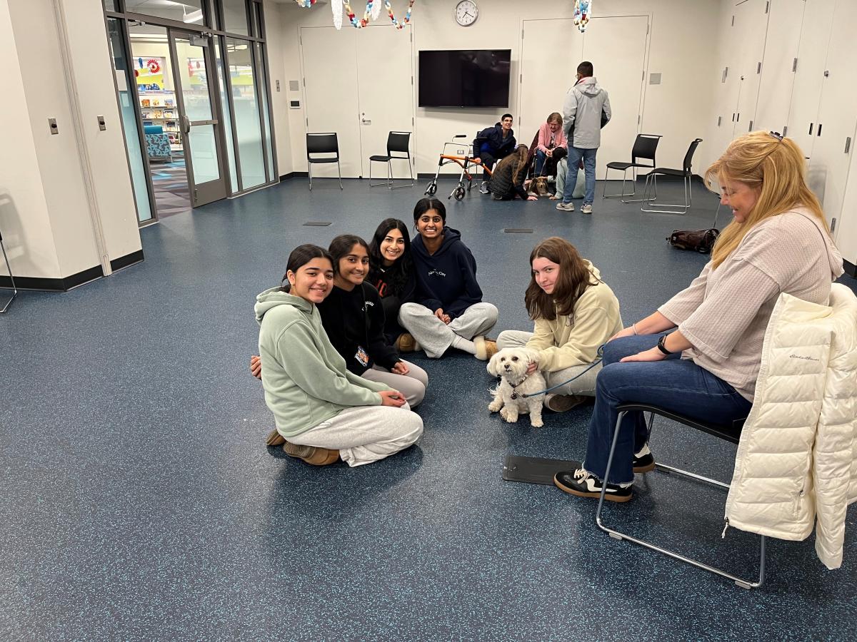 photo of a woman sitting in a chair and 5 teens sitting on the floor surrounding a small white dog.